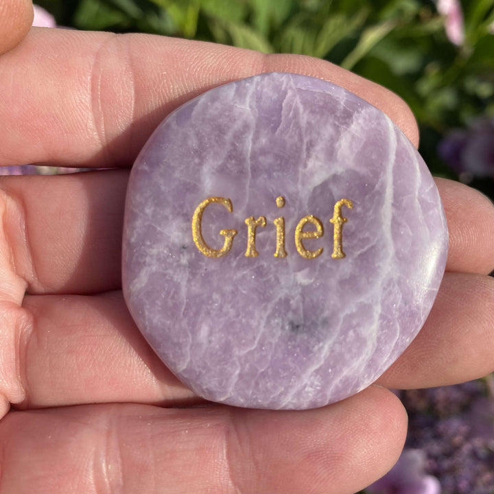 A close-up view of the polished lilac Lepidolite palm stone resting safely in a hand. The smooth, texturised surface and organic details of the engraved crystal are showcased against a soft garden background, capturing peaceful resonance to quieten daily mental chatter and support the Crown Chakra.