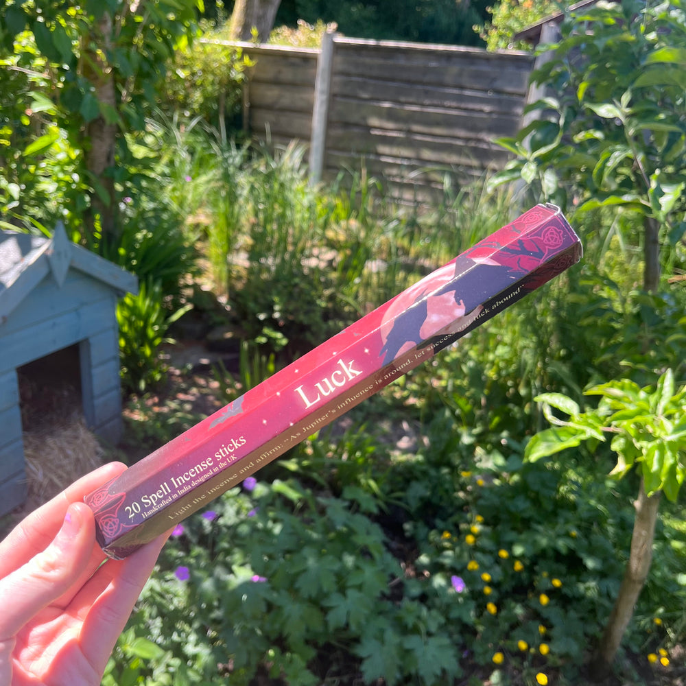 A hand holding the thin pack of luck spell incense sticks at a distance outdoors, highlighting the uplifting floral bouquet fragrance companion styled against sunlit garden greenery to support Crown and Heart Chakra alignment.