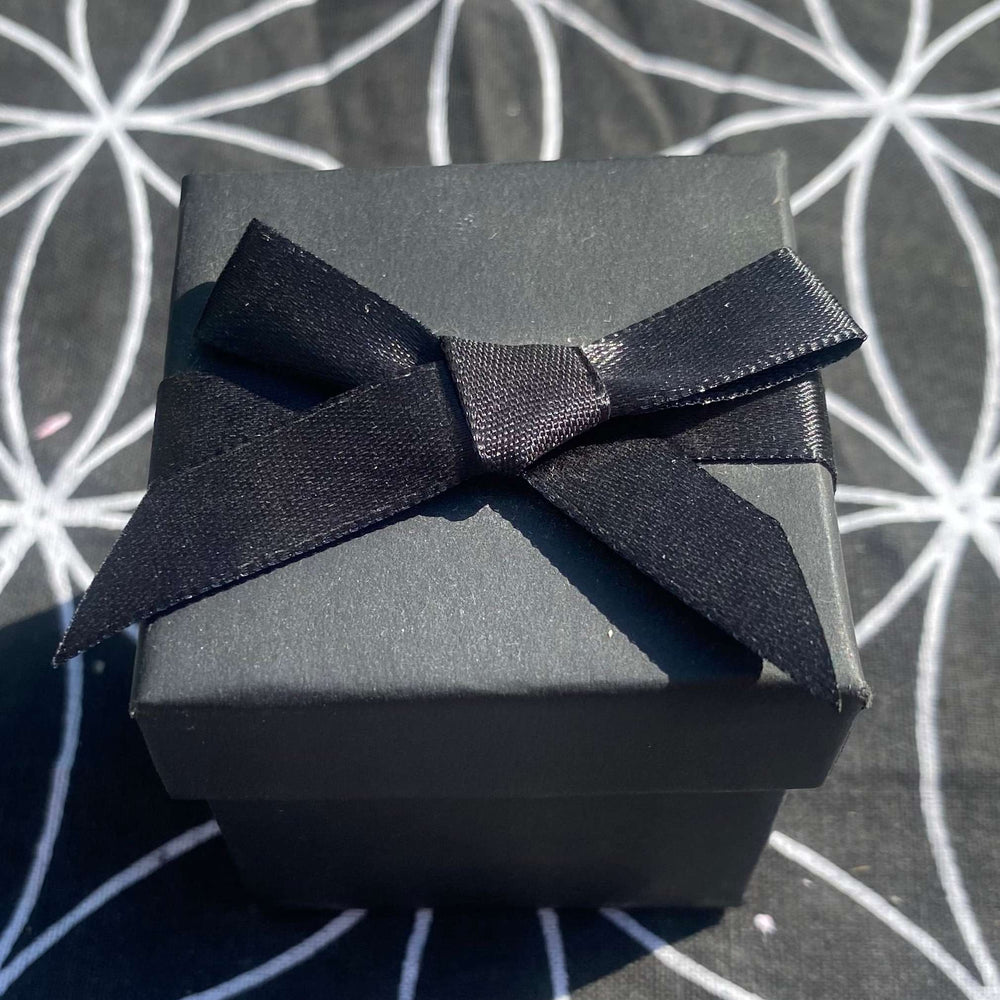A top-down view of the luxury black crystal gift box securely closed with its lid on, resting outdoors on a Flower of Life crystal grid mat. The presentation box is elegantly wrapped with a matching black satin ribbon tied into a neat bow.