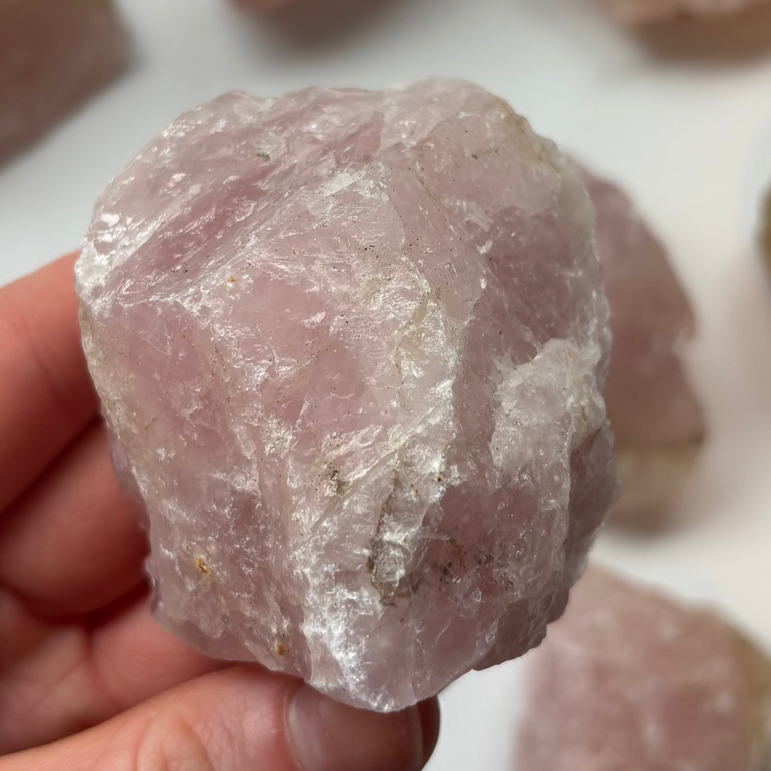 A close-up shot of a medium-sized raw rose quartz crystal chunk held in a hand, showcasing its unpolished texture and soft pink colour layers.