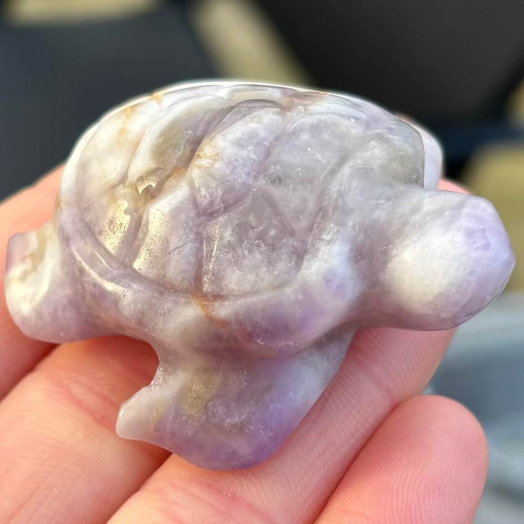 A direct macro close-up shot of a hand-carved Amethyst turtle crystal figurine held flat in an open hand. The polished gemstone creature displays a beautiful blend of natural light purple, lavender, and milky white chevron quartz bands stretching across its smooth, rounded shell structure.