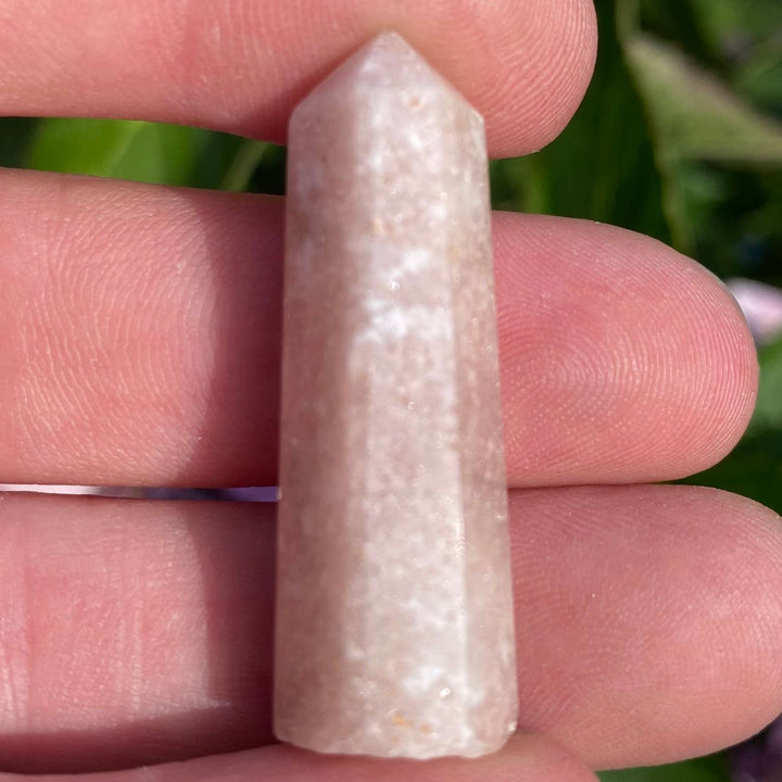 A close-up view of a small, pointed Mini Pink Amethyst Crystal Tower held upright between fingers. The polished generator point showcases soft rose and lavender tones with delicate, frosty mineral pathways, framed by a blurred background of green leaves and pink garden flowers.