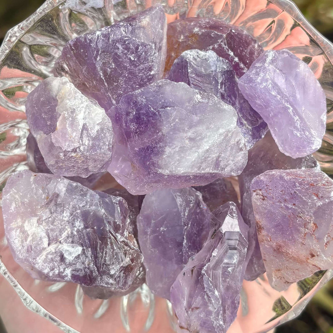 A close-up overhead shot of a glass dish filled with mini raw Amethyst crystal chunks. The natural, unpolished gemstone specimens display irregular, jagged edges and a range of translucent purple tones, from pale lavender to deep violet, highlighting their raw geological structures.