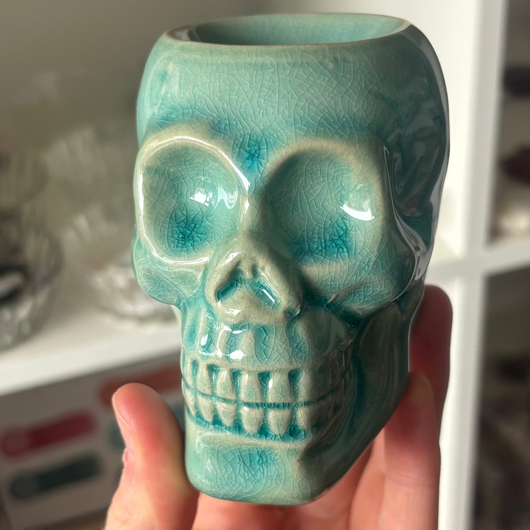 A direct close-up shot of a ceramic mini skull oil burner in a vibrant turquoise blue shade, held gently in a hand. The holder features a glossy glazed finish highlighting its realistic skull features, hollow eye sockets, and clean tooth detailing, set in front of blurred white crystal store shelves.