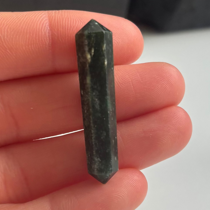 A close-up view of a small, polished Mini Tree Agate double-terminated point crystal held upright between fingers. The double-pointed stone showcases deep forest green and milky white patterns with minor surface scratches, adding unique character to its natural earth journey.