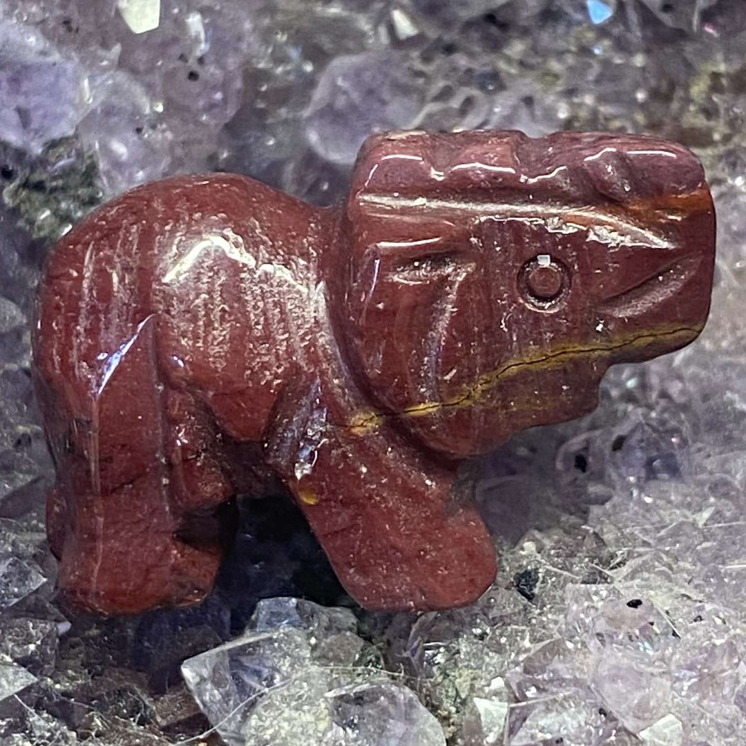 A slightly angled, bird's-eye view of a miniature 3cm carved Mookaite Jasper crystal elephant figurine showcased against a bed of textured raw purple and clear crystal points.