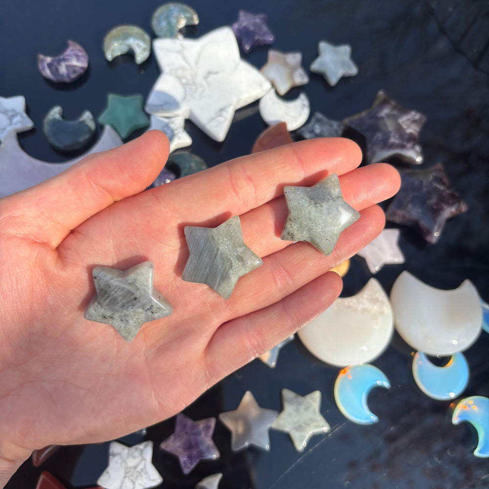 A hand presenting three miniature labradorite star carvings at a distance outdoors over a scattered table layout of moon and star tokens, illustrating the compact 2cm pocket sizing scale of these dreamily magical specimens.
