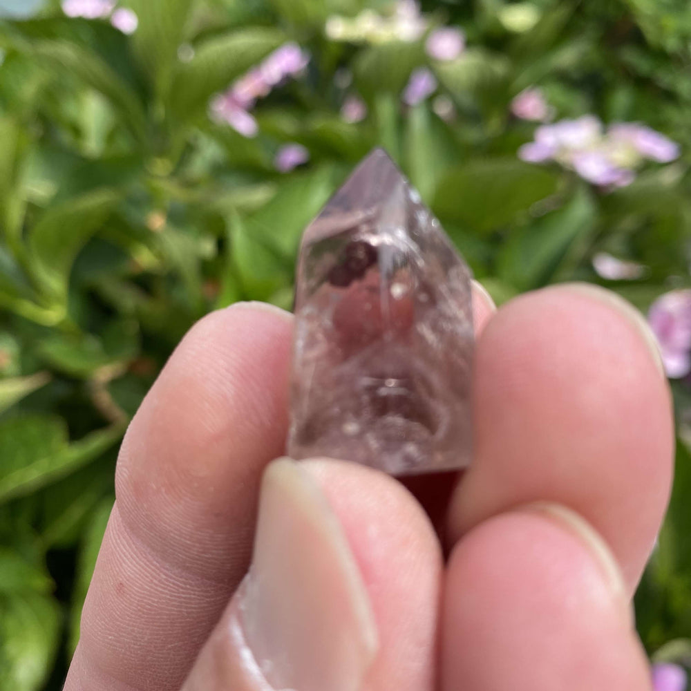 A hand holding a miniature smoky quartz crystal obelisk tower outdoors, highlighting the polished facets and natural internal veils of the stone.
