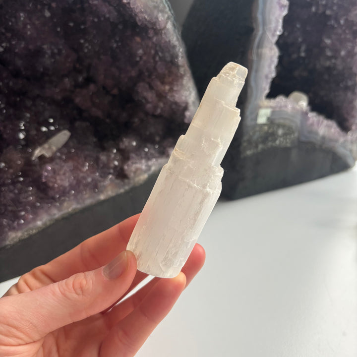 A hand displaying the upper faceted tiers of the misshaped selenite crystal tower close up, demonstrating its compact 3cm width and textured unpolished sides, ideal as a budget-friendly companion to cleanse your gemstone collection.