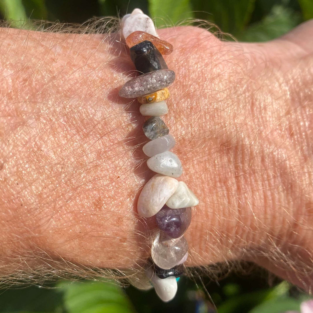 A multi-coloured mixed crystal chip bracelet worn on a wrist outdoors, showcasing an assortment of polished, irregular gemstone beads. The detailed view highlights semi-translucent rose quartz, violet amethyst, tiger eye, and clear quartz chips against a background of green garden leaves.