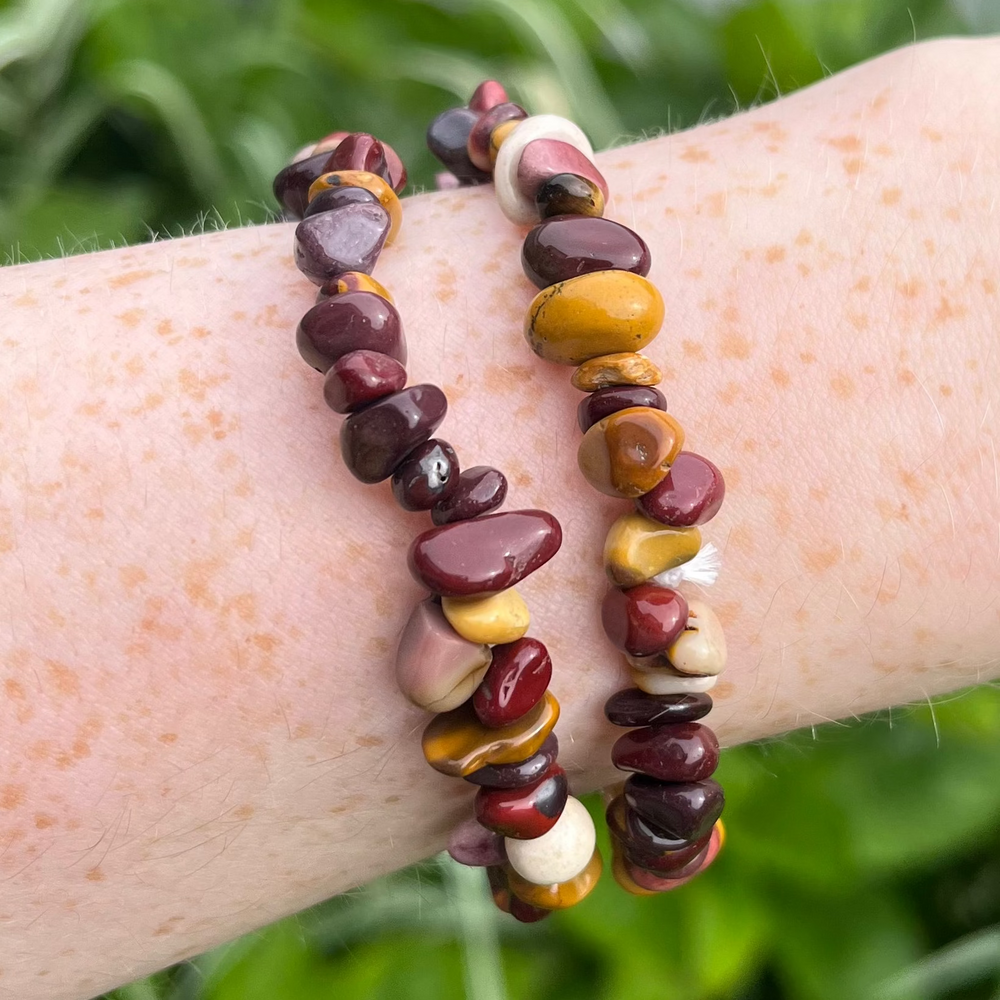 A high-angle shot looking down at two stacked Mookaite crystal bracelets on a wrist. Set against a lush backdrop of vibrant green garden foliage, the image details the comfy fit and unique colour layout of the polished jasper chip stones.