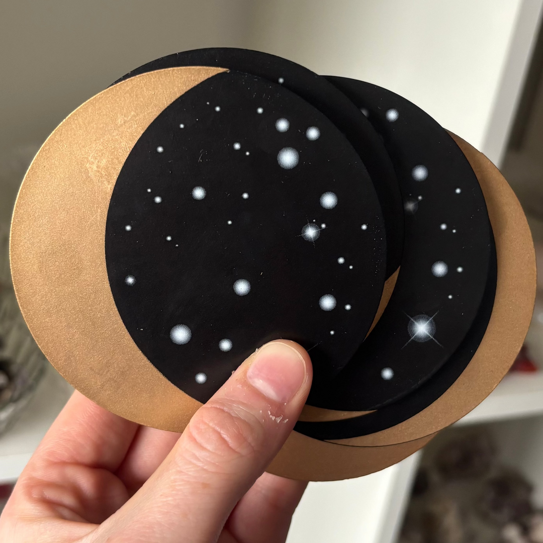 A close-up view of a hand fanning out a set of five circular Moon Phase coasters. The celestial design displays black-and-white starfield patterns layered over natural brown backgrounds, shown indoors to highlight their Third Eye Chakra alignment.