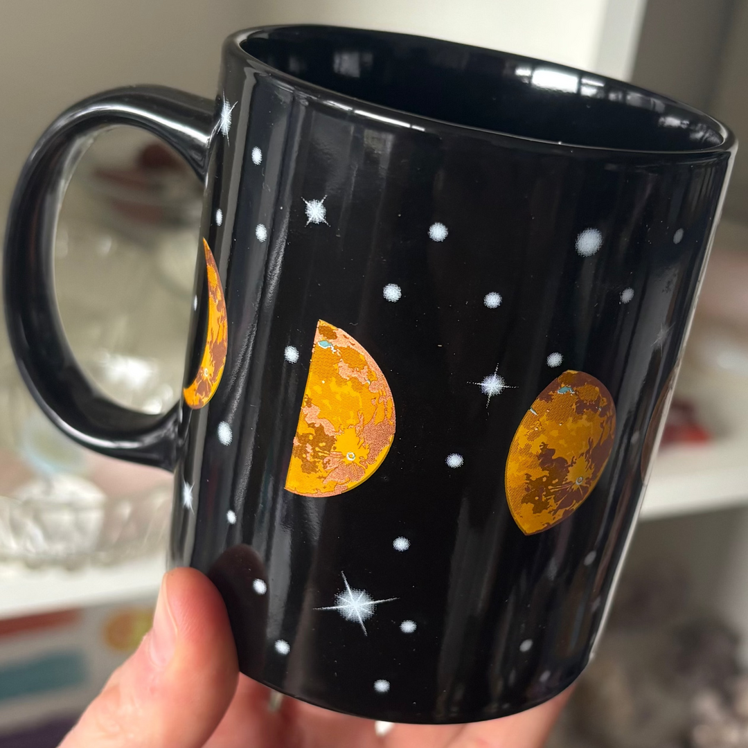 A close-up shot of a glossy black ceramic mug held up in a hand. The exterior features a detailed wrap-around graphic design showing various lunar cycles in warm golden-orange and yellow tones, surrounded by small white star constellations and dot clusters against a dark cosmic background, shown inside a crystal boutique.
