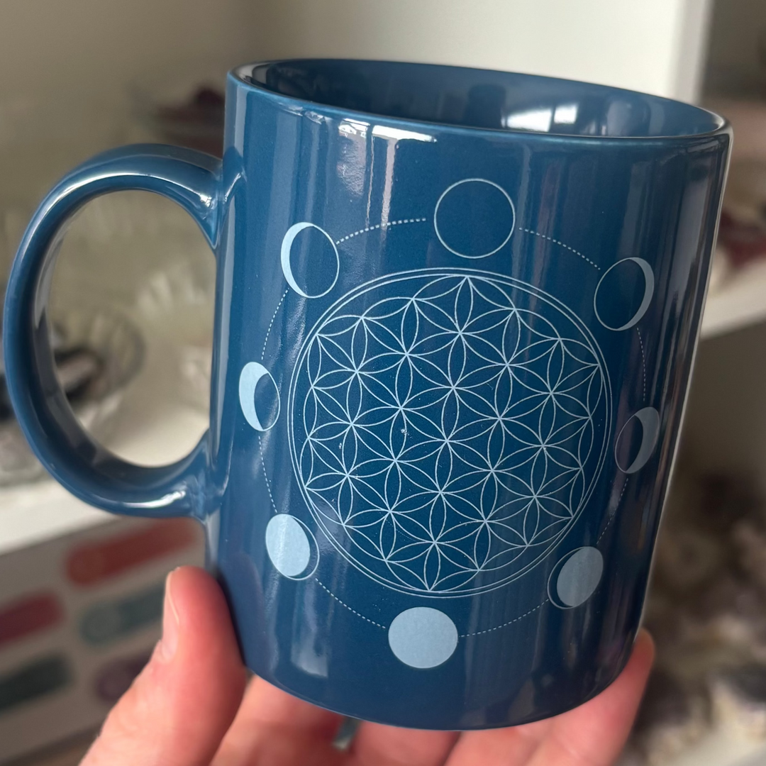 A close-up view of a blue ceramic ritual mug featuring a white geometric flower design held by its handle. The sacred geometry print showcases an intricate Flower of Life motif encircled by lunar phases, beautifully highlighting its Third Eye and Crown Chakra connection.