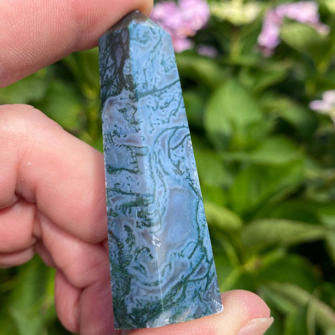 A close-up view of a polished Moss Agate crystal tower held gently between fingers outdoors. The generator point displays beautiful, deep forest-green botanical moss patterns and unique swirls of translucent chalcedony against a blurred garden backdrop.