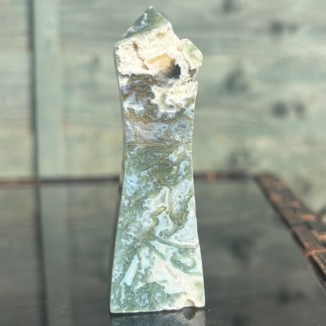 A close-up view of a polished Moss Agate crystal tower standing on a dark reflective surface. The generator point features dense forest-green botanical inclusions layered beneath translucent white milky quartz chalcedony paths to celebrate natural serenity.
