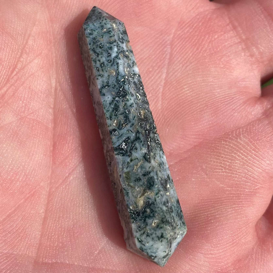 A close-up view of a tiny, double-pointed Moss Agate pocket stone resting diagonally in an open hand outdoors. The slender crystal showcases its dark forest-green botanical patterns and dense, milky quartz mineral layers across its polished surface.