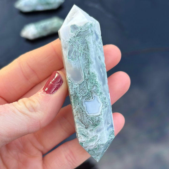 A close-up shot of a polished Moss Agate double terminated crystal point held in a hand, showcasing its vibrant forest-green mossy inclusions and clear quartz layers.