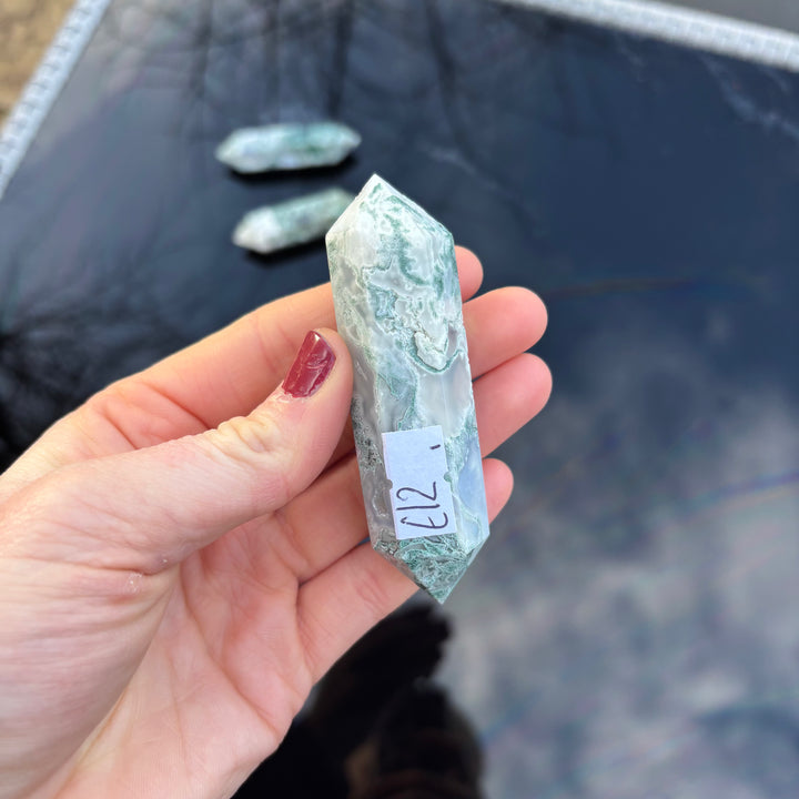 A hand displaying a detailed view of a polished Moss Agate crystal point with unique Earth-born botanical markings and organic textures.