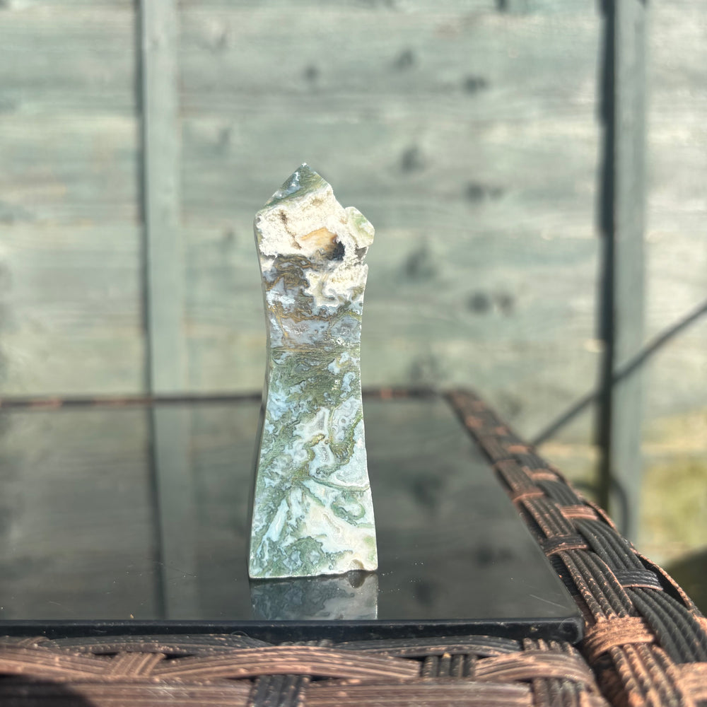 A wide shot looking at the standing Moss Agate crystal generator point arranged on an outdoor table surface. The glass tabletop captures a mirrored reflection of the nine-centimetre gemstone obelisk against a soft, blurred garden backdrop.
