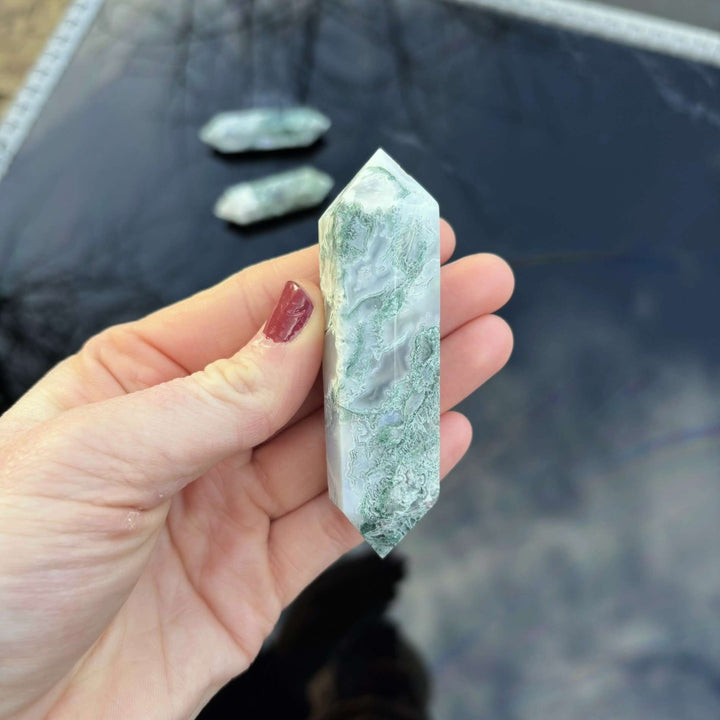 A person holding a multi-faceted Moss Agate double terminated crystal wand, highlighting the deep emerald green mineral swirls inside the stone.