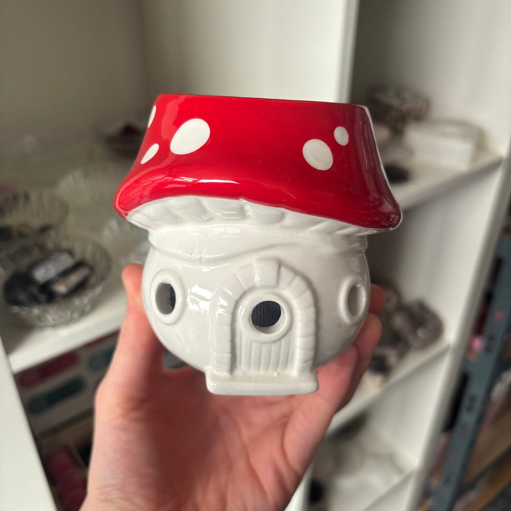 An eye-level shot showing the ceramic toadstool aroma burner held carefully before a light-coloured shop storage shelf. The natural indoor lighting showcases its cute sculpted doorway and window detailing, offering a playful, cosy addition to a bedside table or windowsill sanctuary.