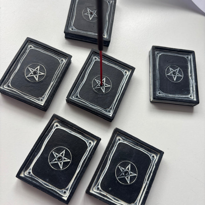 An eye-level shot looking down at several black resin grimoire incense trays grouped together on a white display surface with one holding a burning stick. The sturdy, texturised design serves as a protective ash catcher, bringing a safe harbour and a captivating spiritual aesthetic to any bedside table or altar setup.
