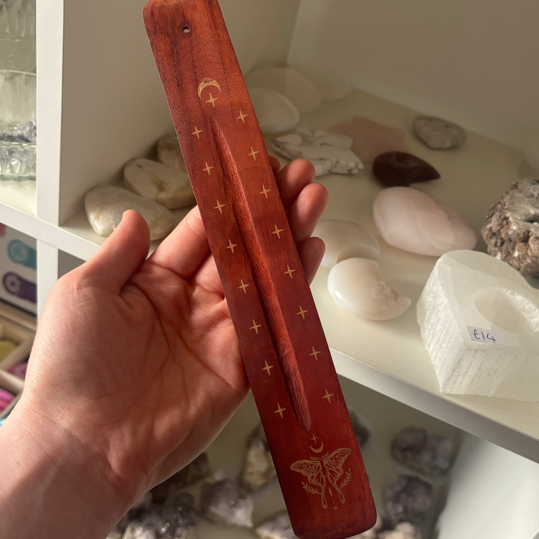 A close-up shot of a long, reddish-brown wooden ash-catching incense ski ski boat or holder held up in a hand. The rustic wooden tray features gold-engraved details including small star symbols along its center groove, a crescent moon motif near the top placement hole, and a highly detailed mystical moth illustration at the base, shown in front of white crystal shop shelves.
