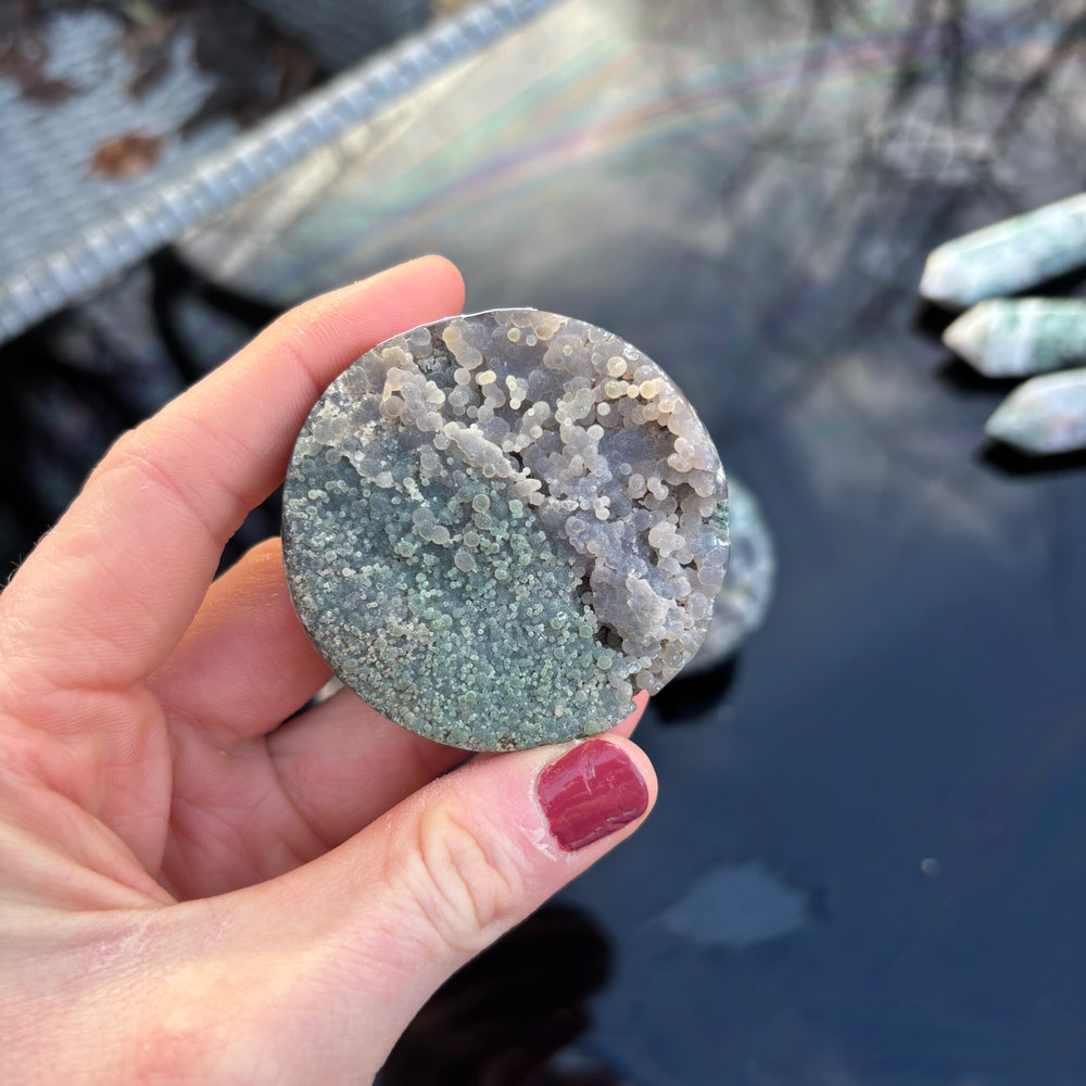 An outdoor shot showing the round purple and green Grape Agate mineral specimen held in a hand from a distance, illustrating its compact pocket scale under daylight.