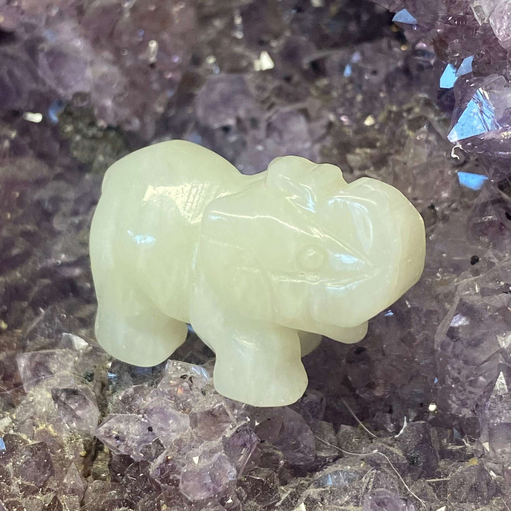 An elevated side-profile view of a small carved jade elephant gemstone figurine showcasing its pale green mineral colors against raw crystal fragments.
