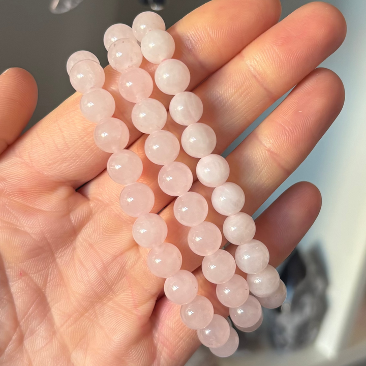 An open hand holding three stacked Rose Quartz round beaded bracelets to demonstrate their styling potential. The layout highlights the uniform bead sizing and the warm, nurturing pink glow of the gemstone pieces.
