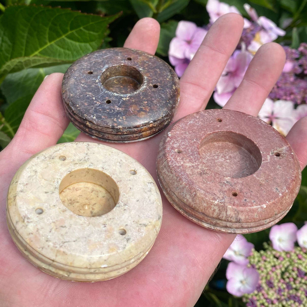 An open hand holding three circular soapstone incense stick and cone burners in a cluster outdoors. The pieces showcase a beautiful variation of natural mineral colourways, including deep charcoal brown, light beige, and a warm terracotta pink.