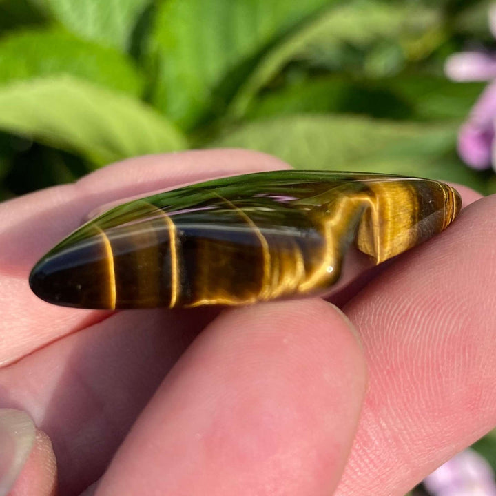 A close-up profile shot showing the rounded, smooth edges of the natural tiger eye celestial carving as it lies across a hand outdoors. The golden-brown bands glimmer softly in the sun, presenting a comforting spiritual token that makes a thoughtful gift for a loved one.