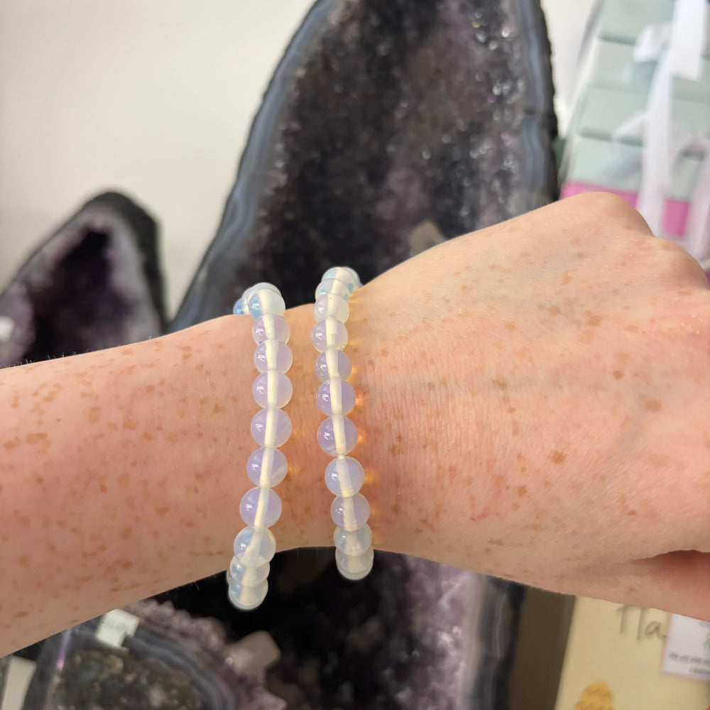 An overhead shot looking down at two stacked Opalite crystal bracelets on a wrist in an indoor shop setting. The lightweight, stretchy bands highlight the uniform shape of the polished round beads, designed to easily bring peaceful clarity to your daily routine.
