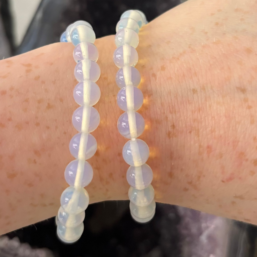A close-up view of two Opalite round beaded bracelets worn stacked on a wrist. The smooth, translucent opalised glass beads showcase a luminous milky blue glow and subtle golden undertones, channelling high vibes and a comforting everyday texture.
