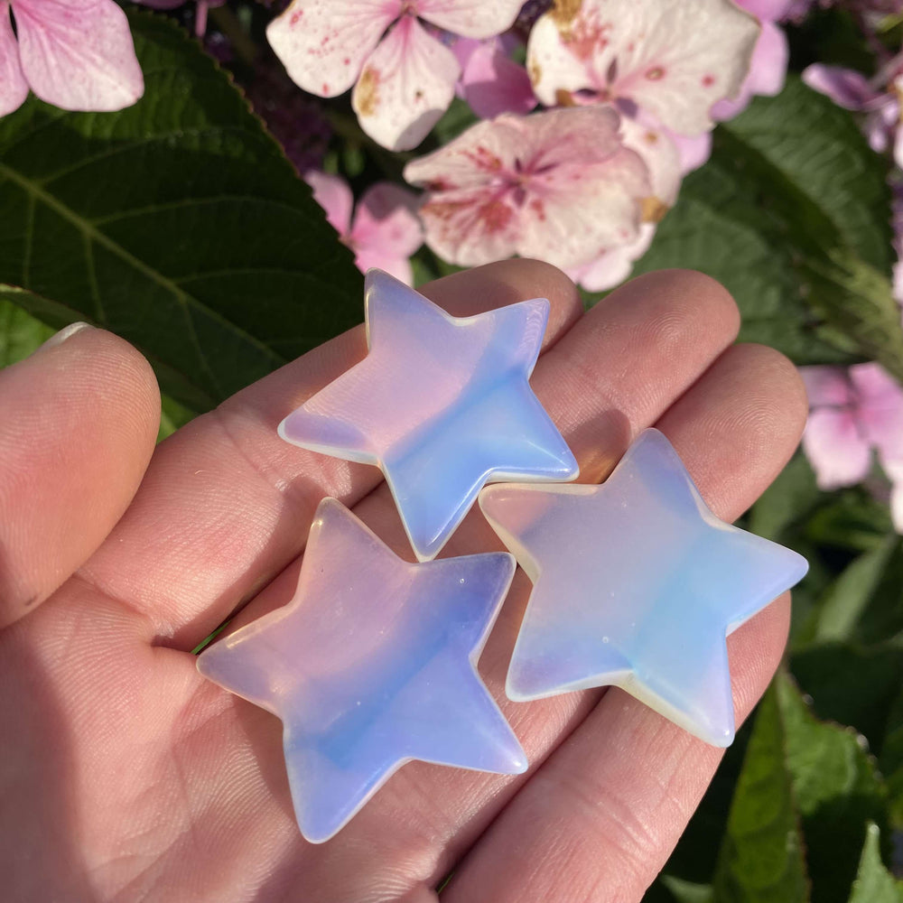 An overhead shot looking down at three iridescent Opalite stars held carefully in an open hand outdoors. Shifting with dreamy pastel blue hues, the smooth, polished edges of these light-coloured pocket companions glow under the morning sun to create a cosy, comforting vibe.