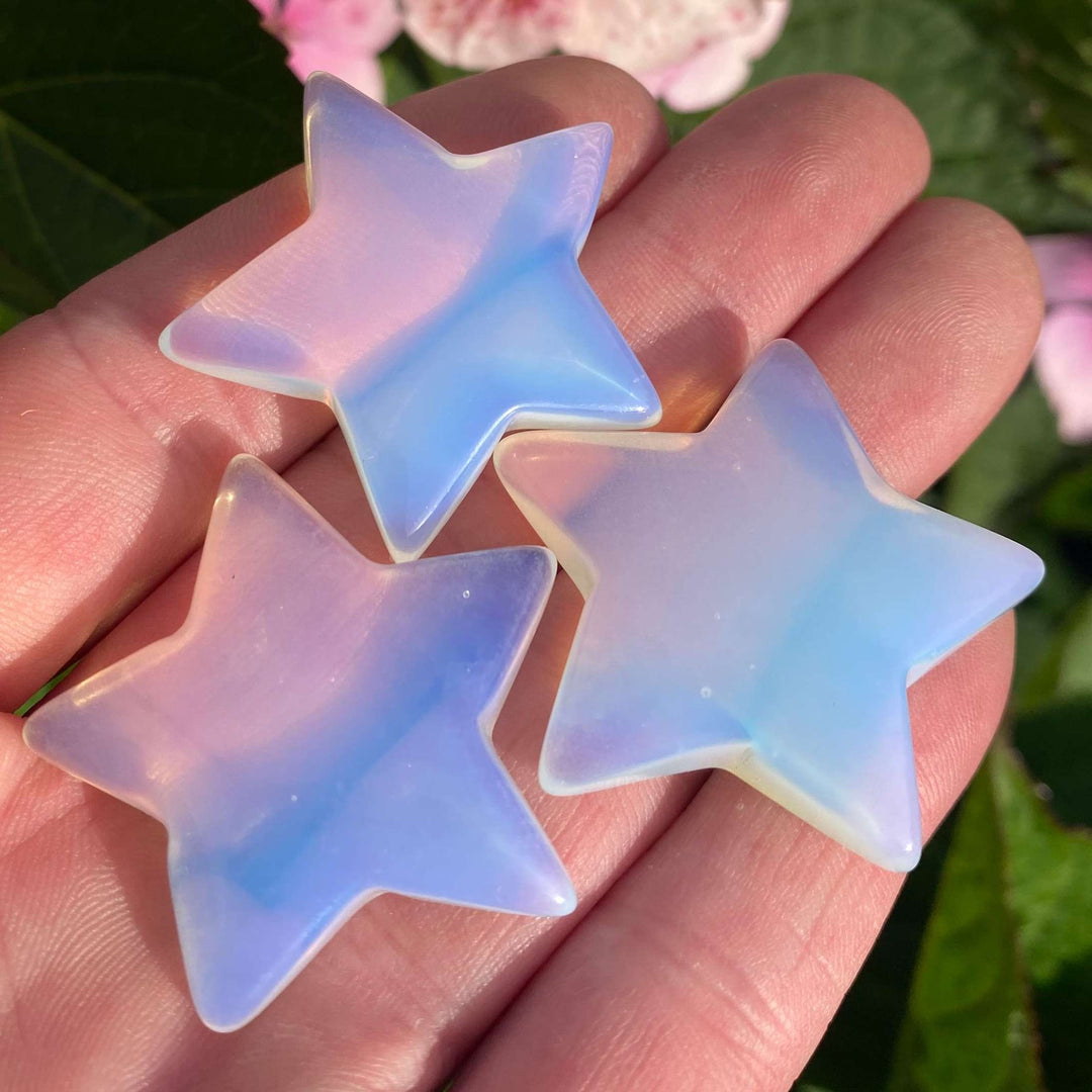 A close-up view of three polished, star-shaped Opalite crystal carvings resting together across an open palm. The milky translucent stones display a soft blue and lavender iridescent sheen against a blurred background of pink garden flowers to highlight their Throat Chakra alignment.