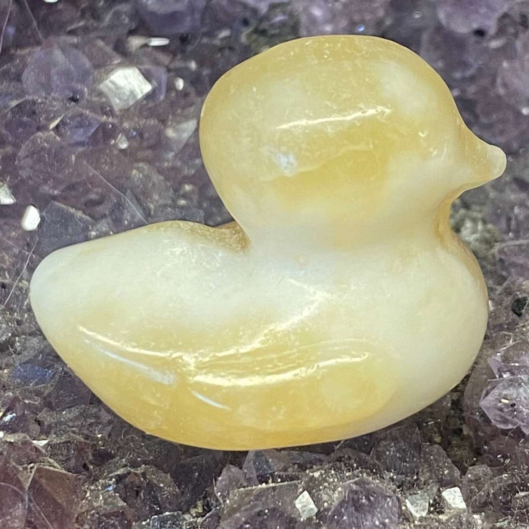 A close-up view of a hand-carved orange calcite duck crystal, showing its smooth, satiny texture and pale honey-yellow to sunset-hued tones. The polished bird figurine is cradled on a bed of purple crystal clusters, radiating themes of warm joy and gentle uplifting energy.