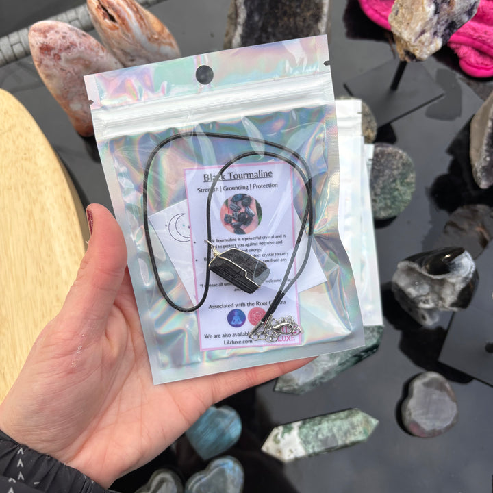 A hand holding the black tourmaline necklace sealed inside its retail cellophane pouch alongside and crystal meaning card, showcasing the gift-ready presentation designed to shield personal energy fields.