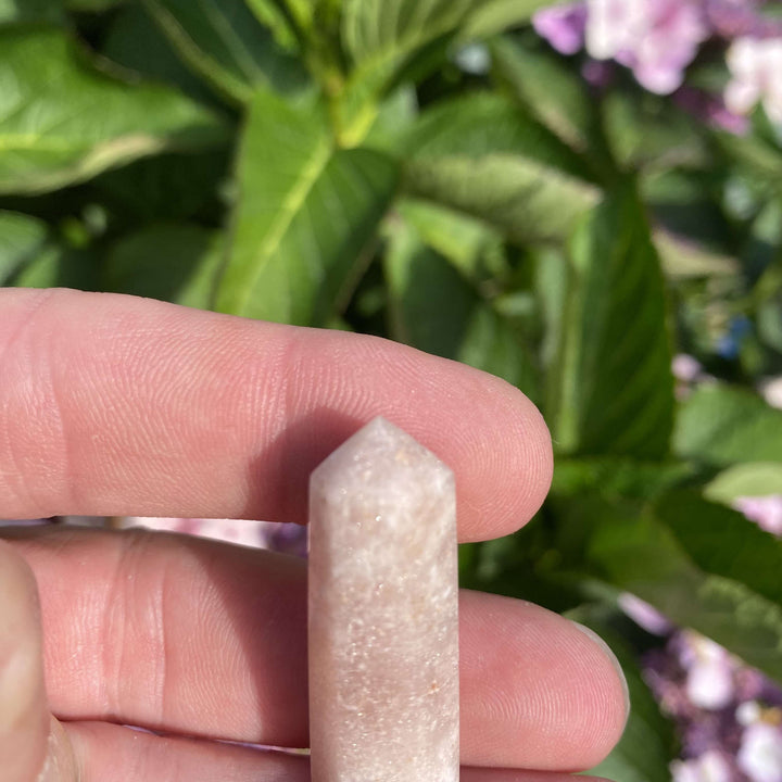 A close-up view of the miniature pink amethyst point angled gently on fingertips before green garden foliage. Its solid, substantial texture and comforting rose hues serve as a graceful token of inner peace, welcoming heart-centred clarity and quiet relaxation into your room.