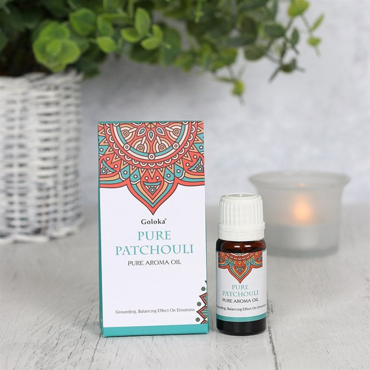 A 10ml amber glass bottle of Pure Patchouli Home Aroma Oil standing next to its white and teal card box featuring an intricate red mandala design, styled on a wooden surface with a lit frosted glass candle and green foliage in the background.