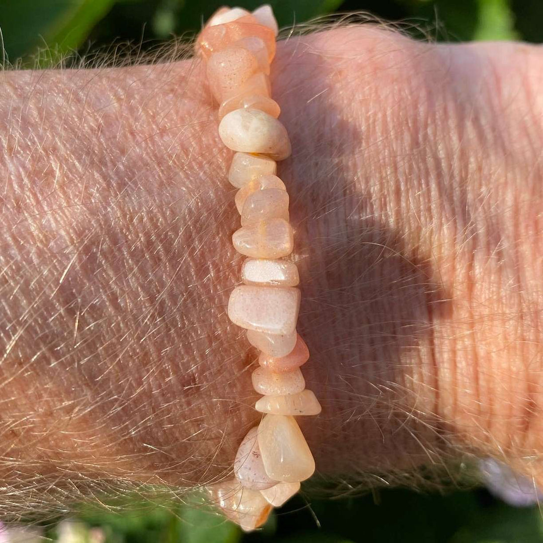 A direct vertical close-up shot of a person's wrist wearing the Peach Moonstone crystal chip bracelet. The polished, irregular gemstone fragments glow with warm peach, salmon, and milky orange tones under bright, natural sunlight, contrasted against a soft-focus green garden background.
