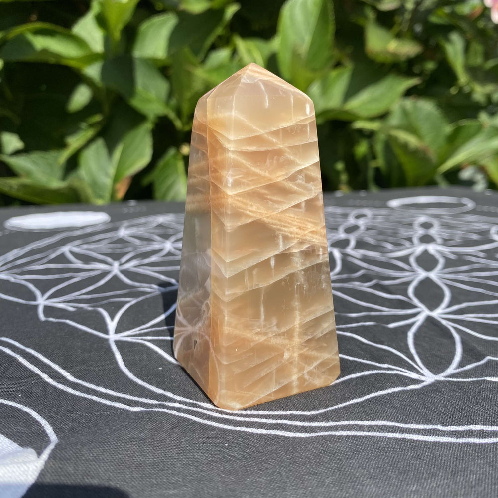 A standalone shot of a faceted Peach Moonstone crystal point standing upright on a black decorative crystal grid mat with geometric patterns.
