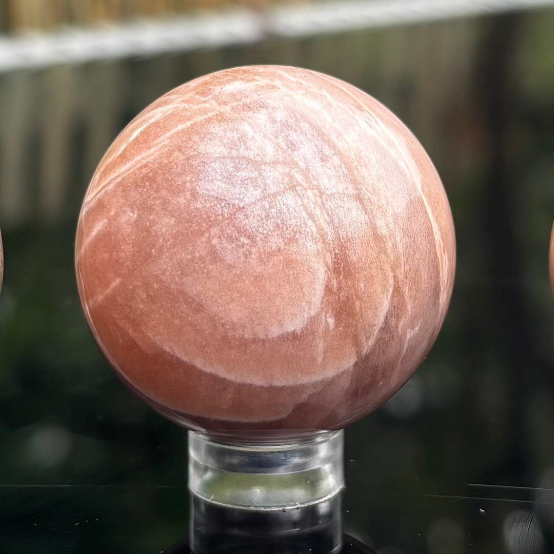 A polished Peach Moonstone crystal sphere resting elegantly on a small, clear acrylic display holder, showcasing its smooth glassy surface, warm peach-apricot colour tones, and curving white mineral veins.