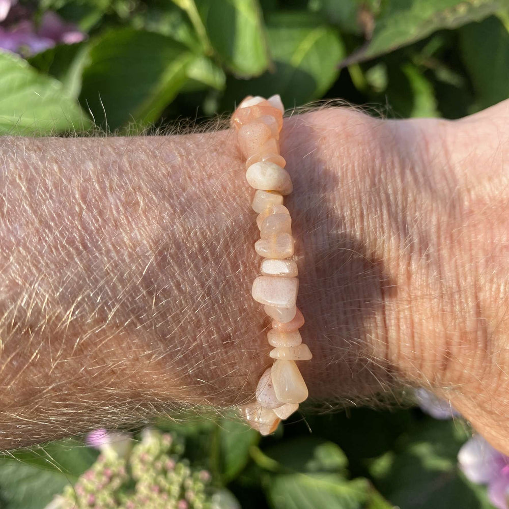 A side-angle outdoor view of the stretchy crystal bracelet worn on a wrist. The individual, soft peach moonstone chips catch the light, showing off their natural, cloudy translucency against a textured background of green garden leaves.