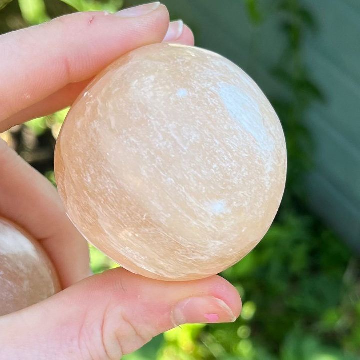 A close-up view of a polished Peach Selenite crystal sphere held between a thumb and fingers outdoors, showcasing its smooth, satiny surface, soft peach-orange colour tones, and fine shimmering fibrous mineral lines.
