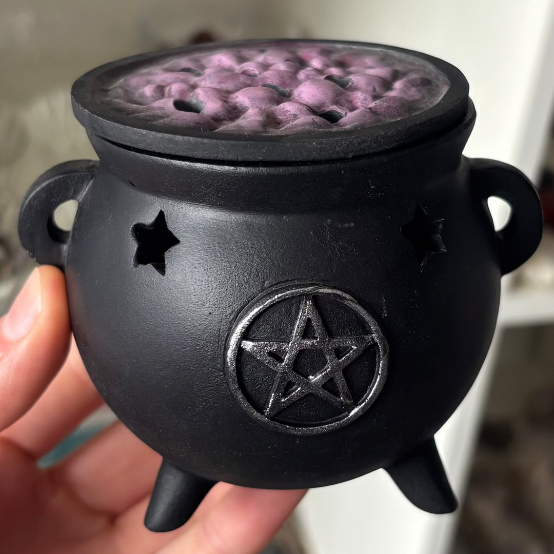 A close-up view of an enchanting resin cauldron incense cone burner held upright in a hand. The matte black cauldron features a distressed silver effect triple pentagram emblem on its face, mystical cut-out star vents, and a removable purple bubbling brew lid designed to let aromatic smoke rise gently.