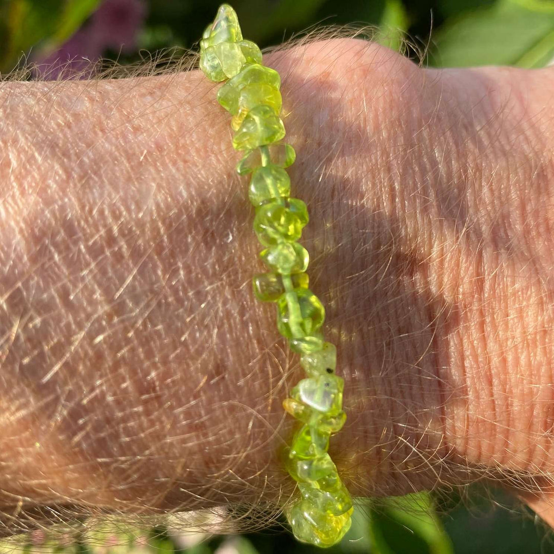A direct vertical close-up shot of a person's wrist wearing a petite Peridot crystal chip bracelet. The tiny, polished gemstone fragments glow with vibrant lime, olive, and translucent chartreuse-green tones under bright, natural sunlight, contrasted against a soft-focus green garden background.