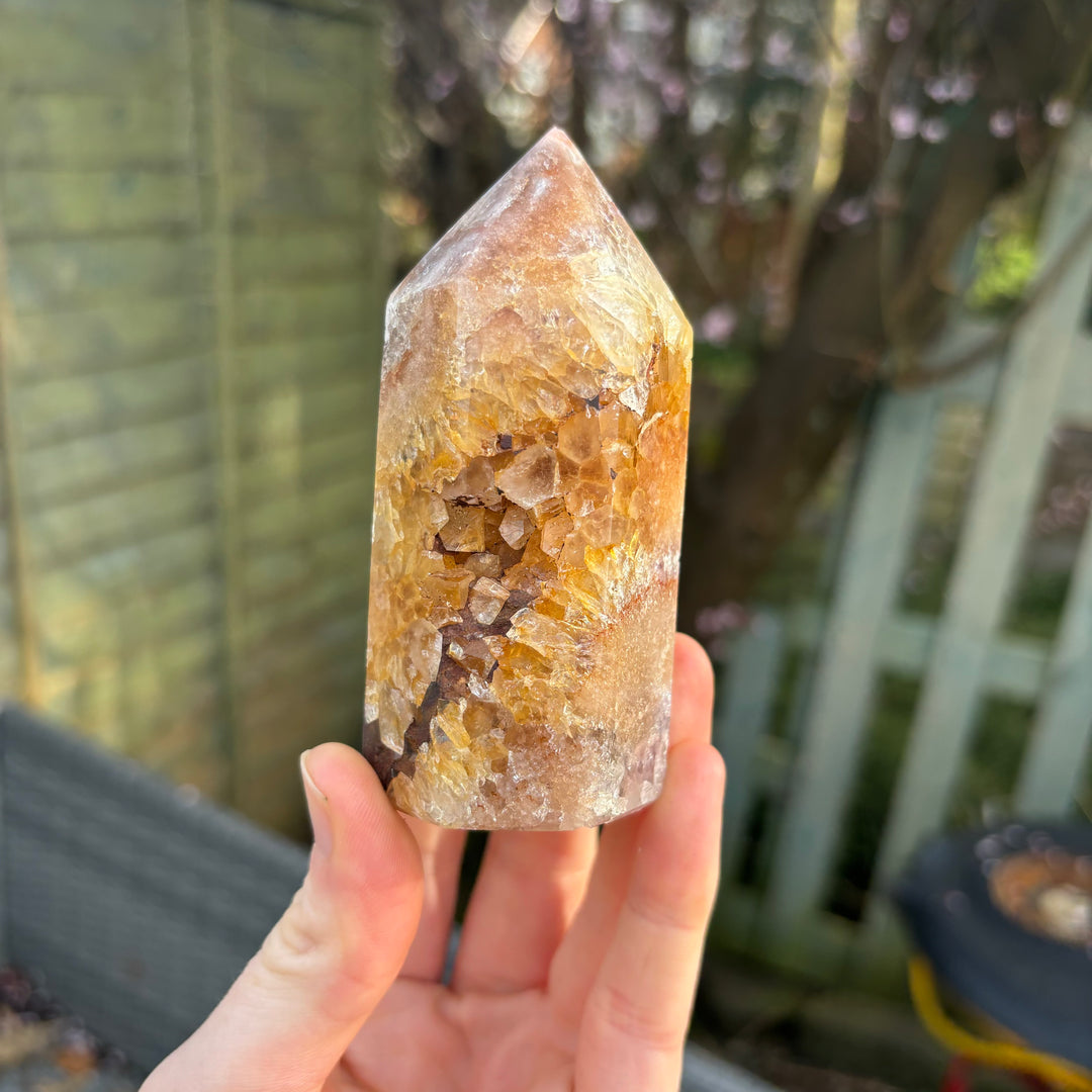 A hand holding the pink amethyst generator point vertically outdoors, demonstrating its approximate 9cm height, 4cm width, and substantial geometric form against a blurred garden fence background.