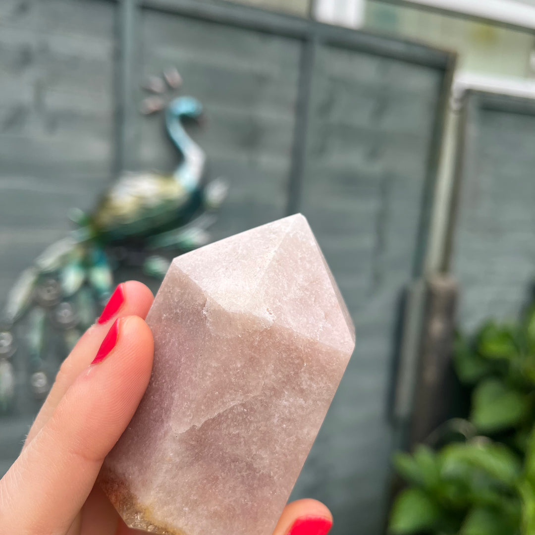 A close-up view of the Pink Amethyst crystal tower angled softly between fingertips with painted nails. The smooth, polished surfaces and heavy mineral textures are fully captured, making it a supportive companion to encourage emotional balance, grace, and inner peace.
